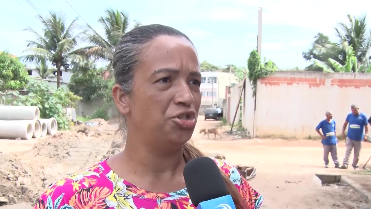 Moradora fala à reportagem, com trabalhadores e manilhas ao fundo
